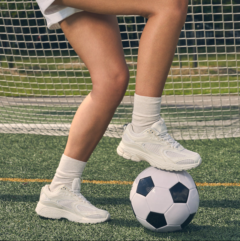 A person in white athletic shorts and white sneakers kicks a black-and-white soccer ball on a green turf field in front of a white goal net.