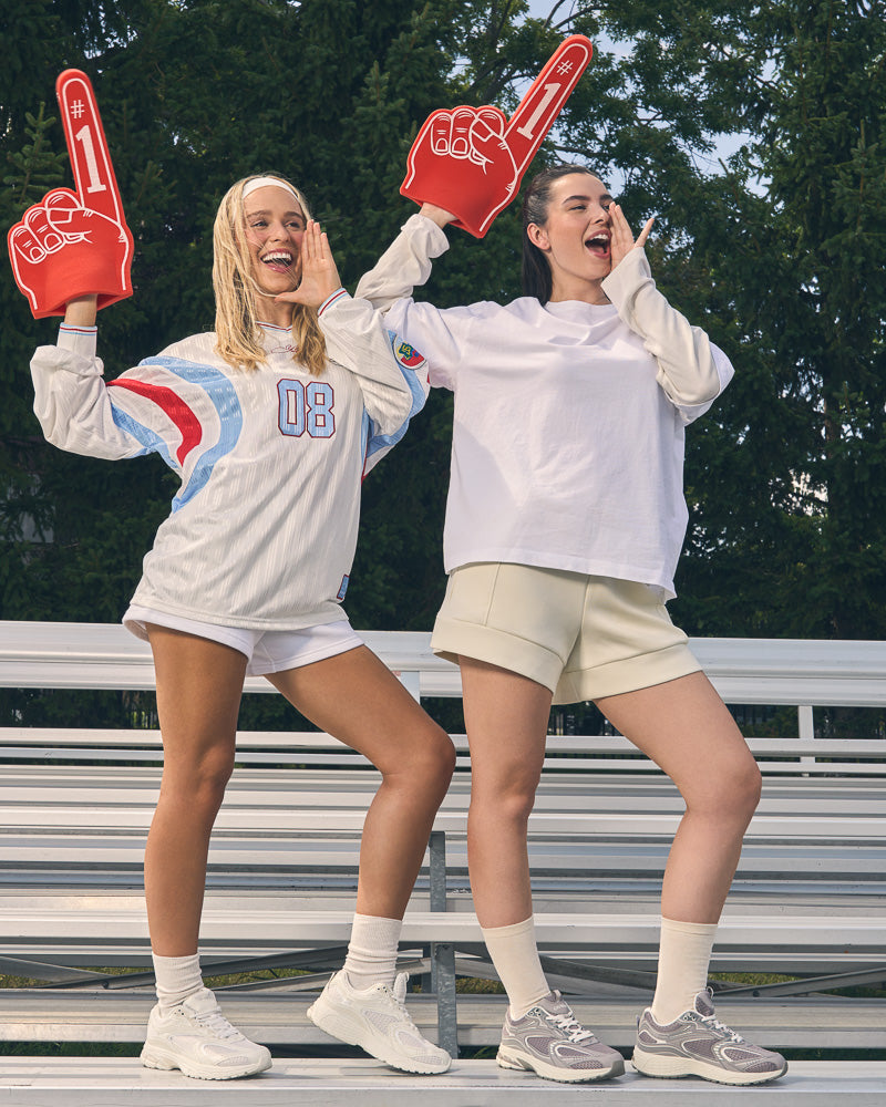Two women standing on outdoor bleachers wearing oversized white sweatshirts, shorts, and Yellow Shoes CHARGE sneakers, each holding a red foam finger. Trees and bleachers are visible in the background.